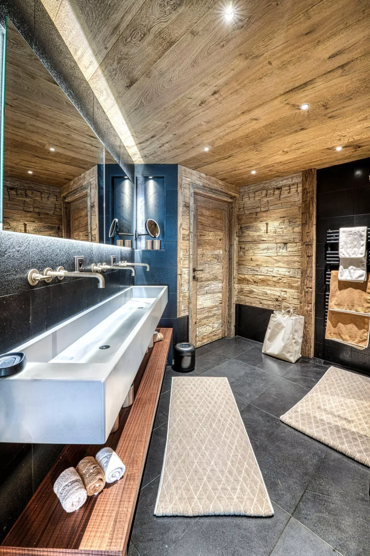 A modern bathroom with a long white sink, dark tiled floor, and wood-paneled walls.