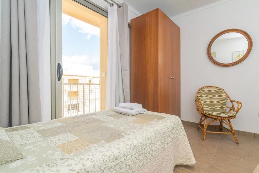 Un dormitorio con una cama, un armario alto de madera y una silla junto a la ventana.