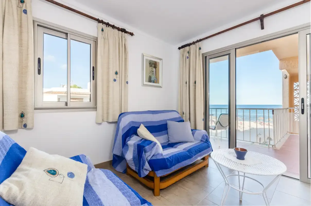 Una sala de estar luminosa con asientos azules, una mesa blanca y un balcón con vistas al mar.