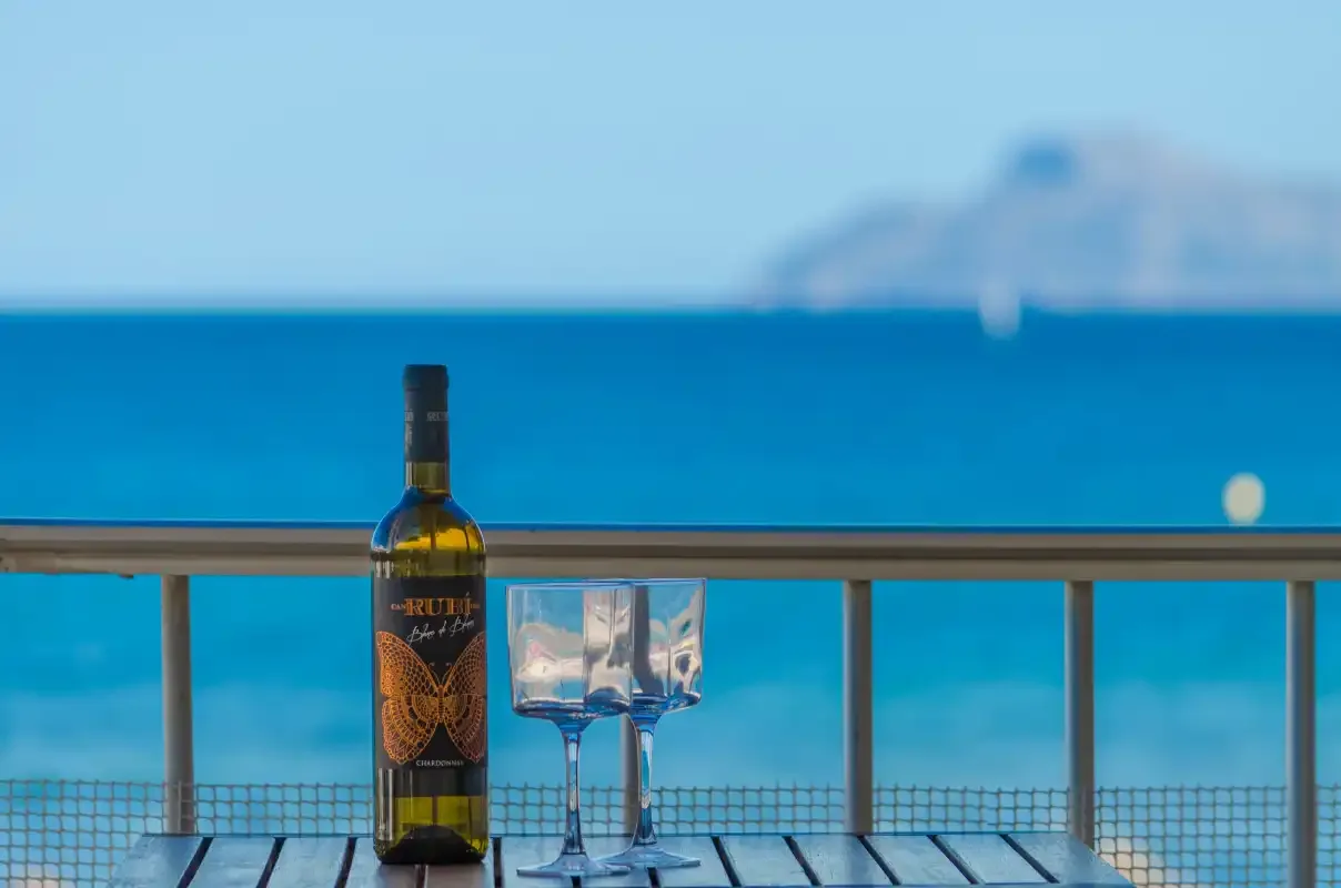 Una botella y unas copas están sobre la barandilla de un balcón frente al mar.