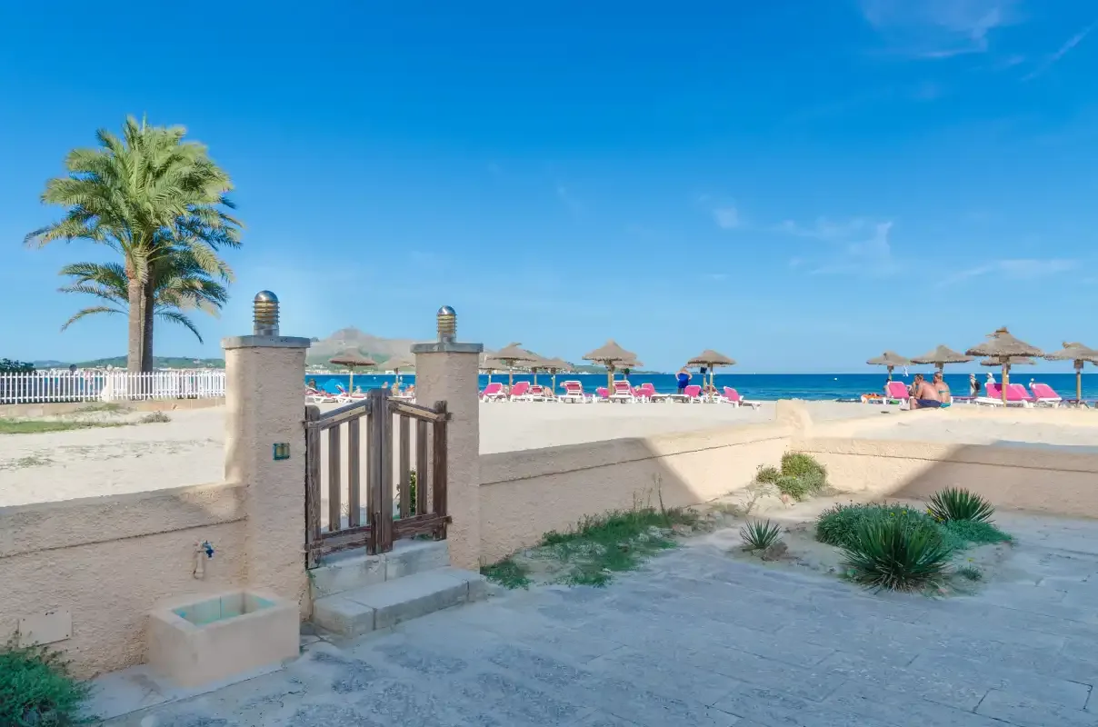Acceso a la playa con una pequeña puerta, una palmera y sombrillas junto a la orilla.