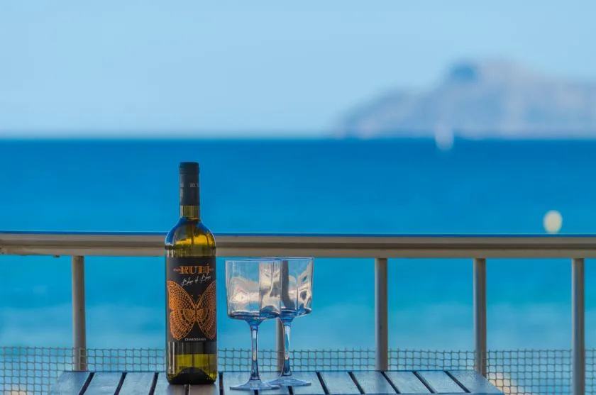 Una botella y dos copas están sobre una barandilla de balcón frente al mar.