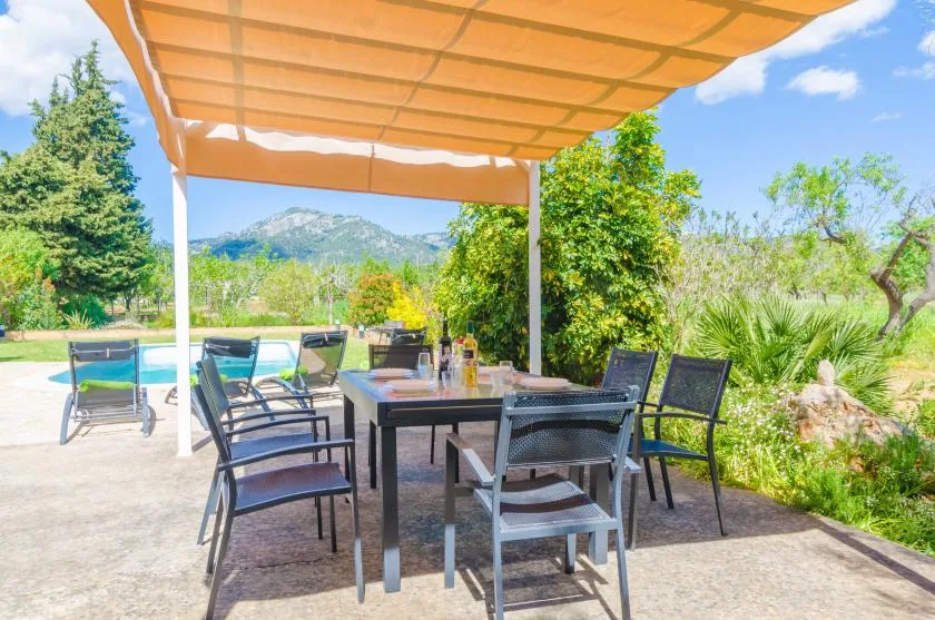 A shaded outdoor dining area with a table and seating overlooks a garden and pool.
