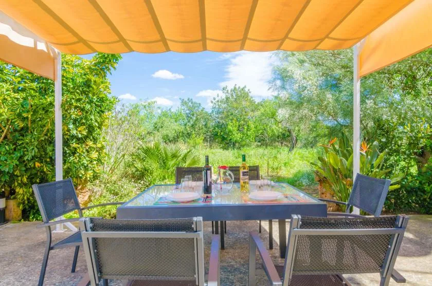 An outdoor dining table with chairs sits beneath an orange canopy amid greenery.