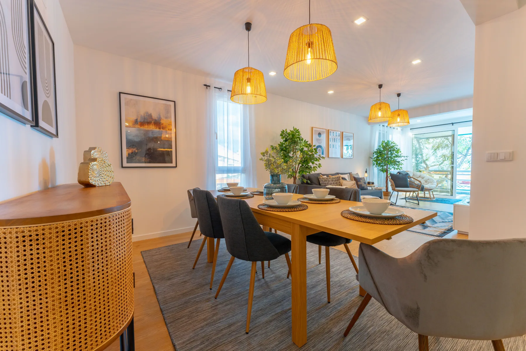 A bright dining and living area features a wooden table and large windows.
