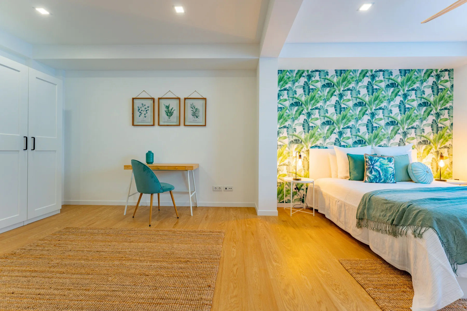 A bedroom with a bed, a small desk, and leafy wall decor.