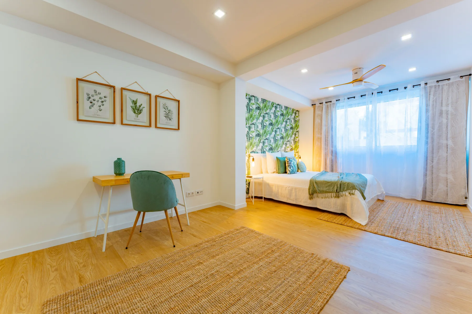 A bright bedroom with a bed beside large curtained windows and a small desk.