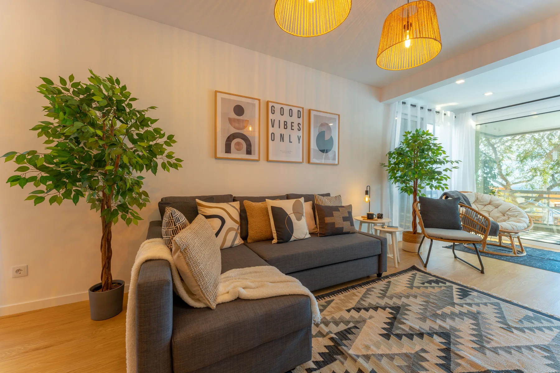 A living room with a sectional sofa, patterned rug, plants, and a seating area by the windows.