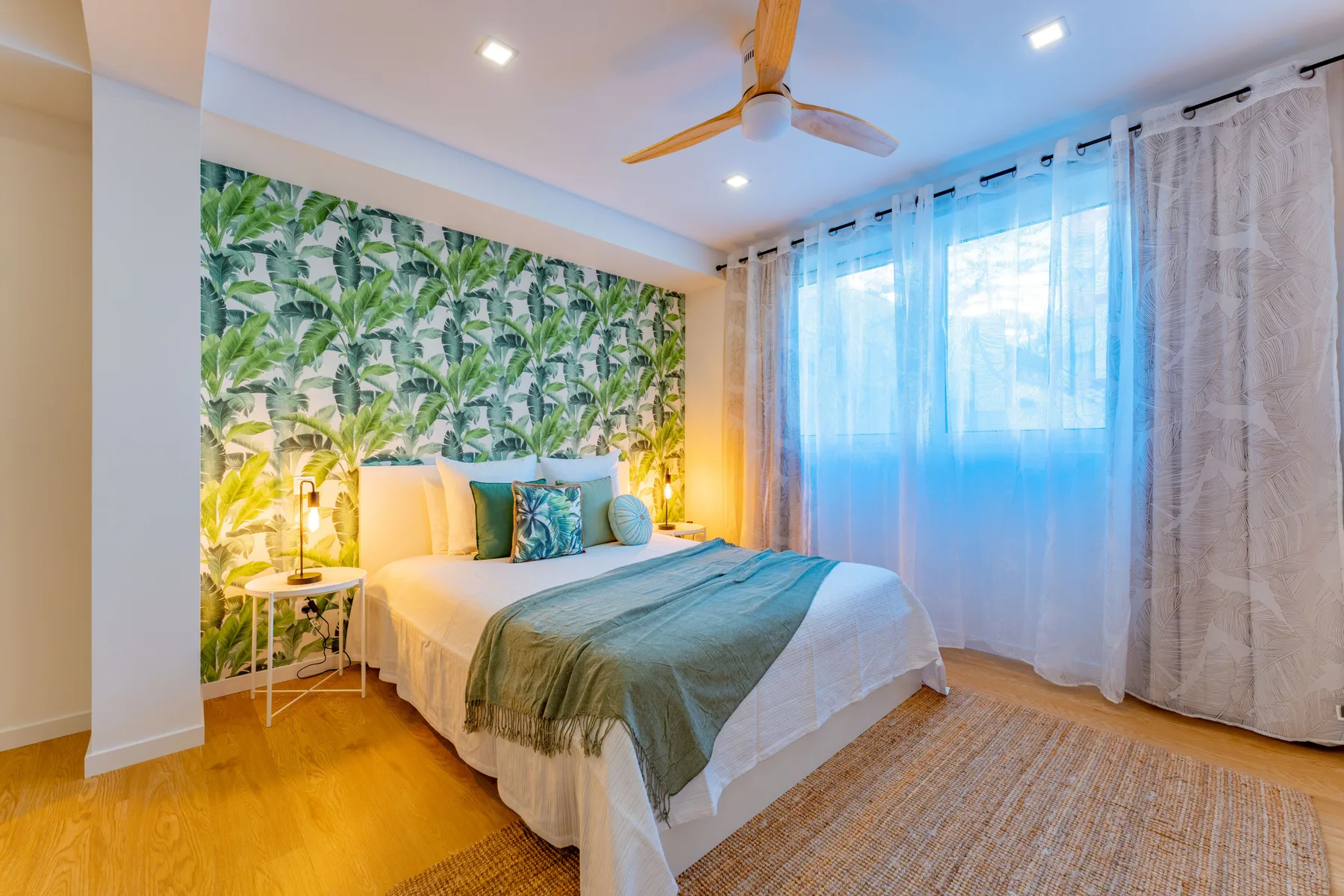 A bedroom with a bed, leafy wallpaper, and sheer curtains.