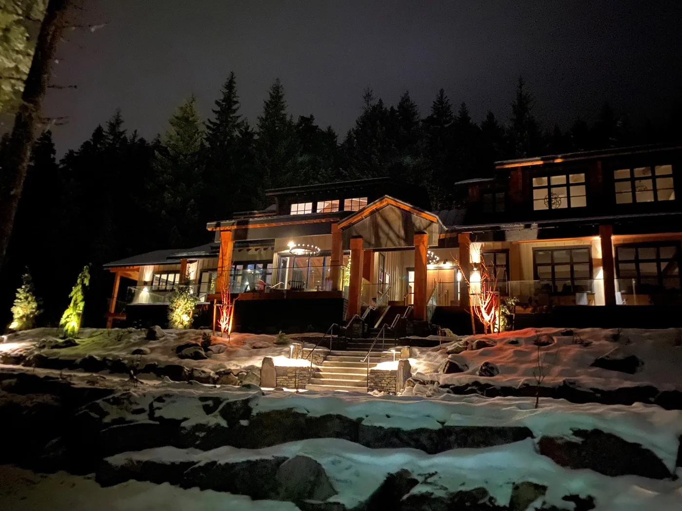 Una casa grande brilla con luces cálidas en una noche nevada y arbolada.