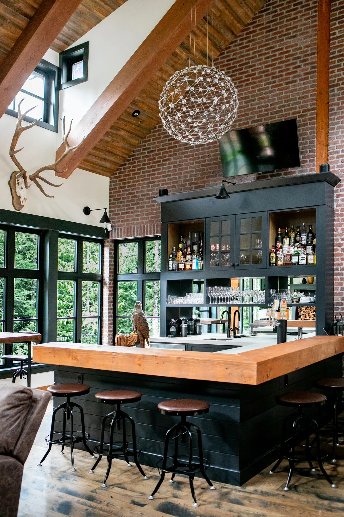 Una zona de bar con taburetes, estantes de botellas y grandes ventanas bajo un techo de madera inclinado.