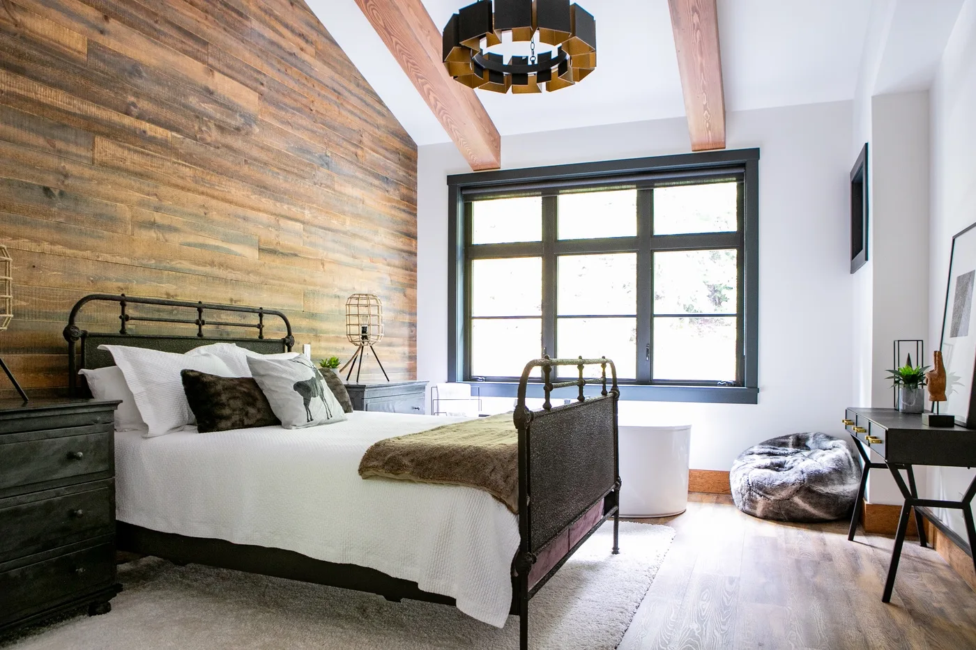 Un dormitorio con una cama de metal, una pared revestida de madera y ventanales.