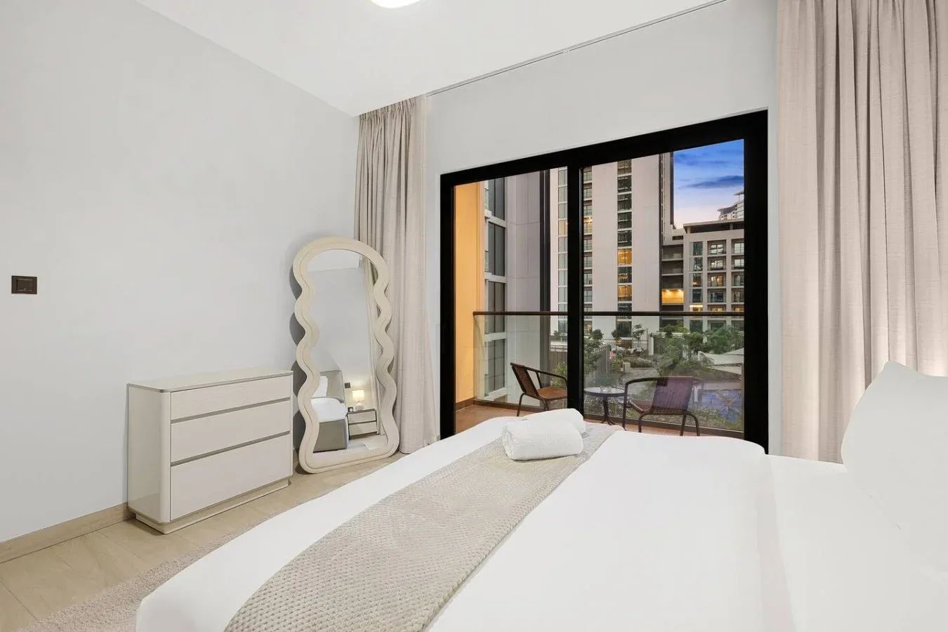 Un dormitorio luminoso con cama, cómoda y una gran ventana con vista a edificios cercanos.