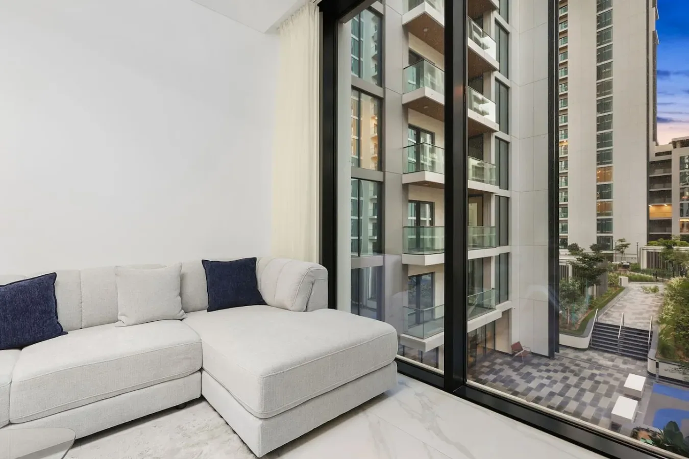 Una sala de estar con asientos claros junto a ventanas amplias con vista a edificios cercanos.
