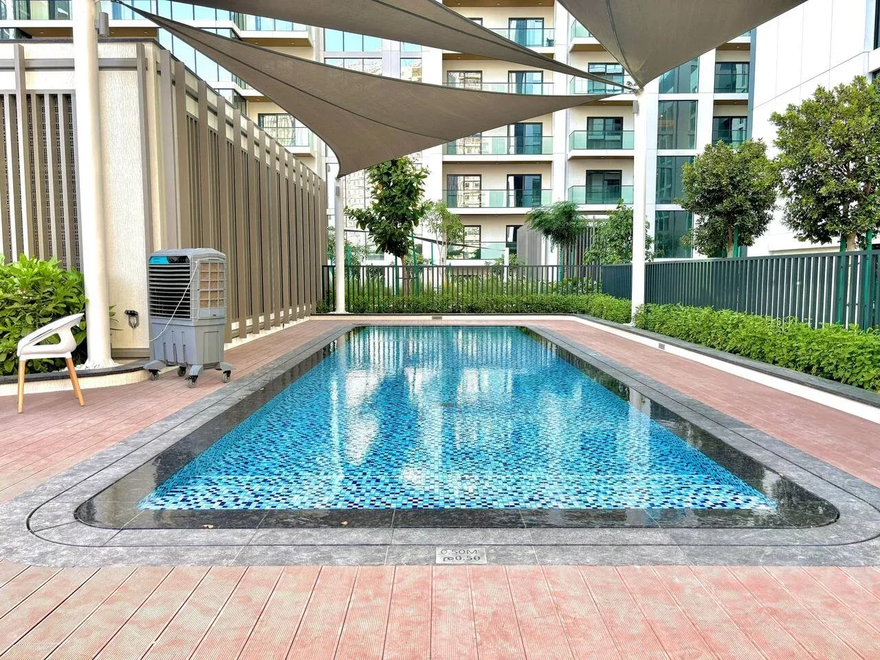 Una piscina al aire libre está bajo toldos de sombra, con edificios de apartamentos al fondo.