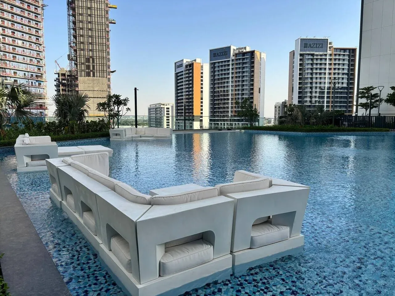 Una piscina en la azotea con asientos blancos junto al agua y edificios altos al fondo.