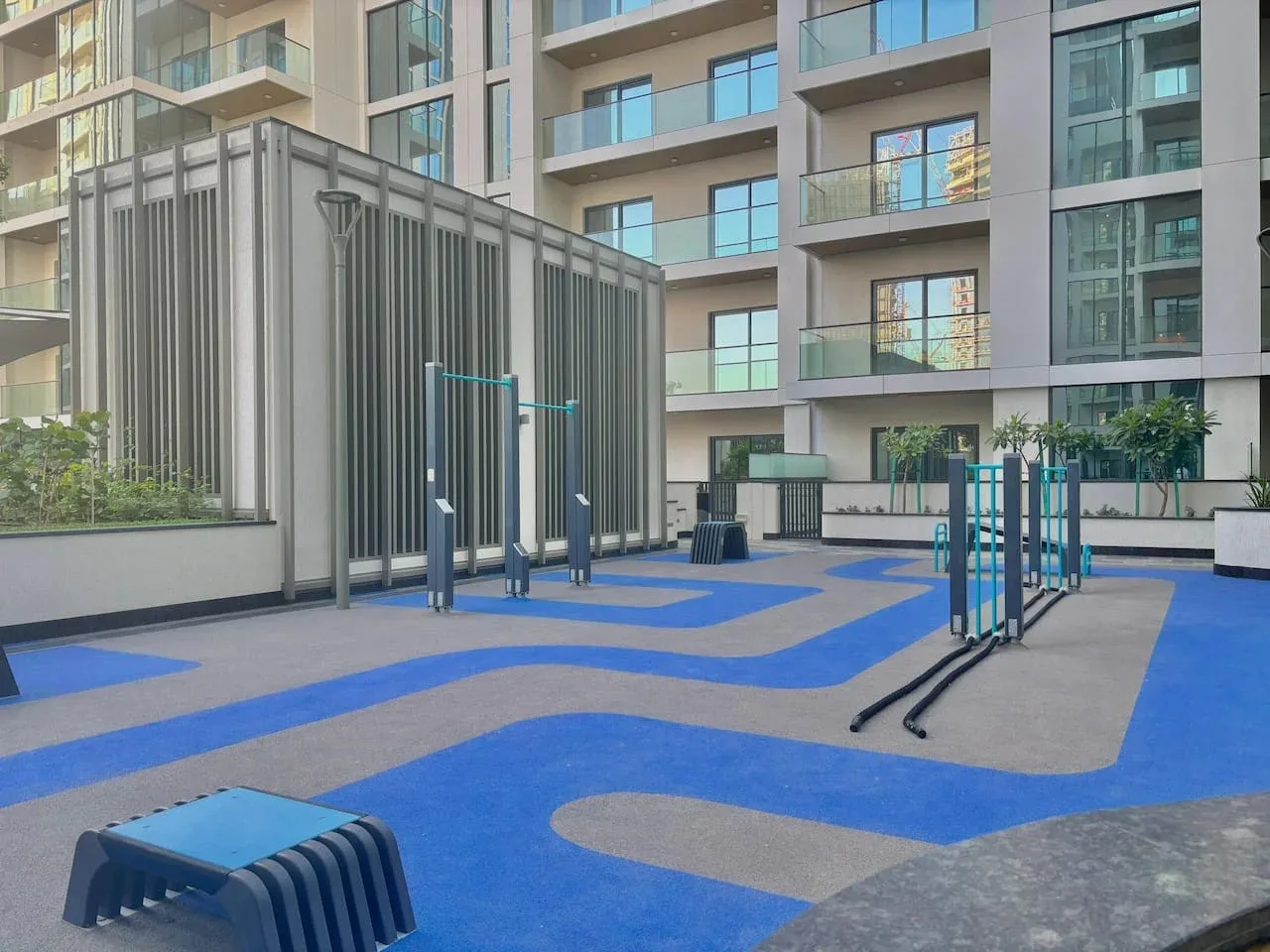 Una zona de ejercicio al aire libre tiene suelo azul y gris y equipos junto a un edificio de apartamentos.