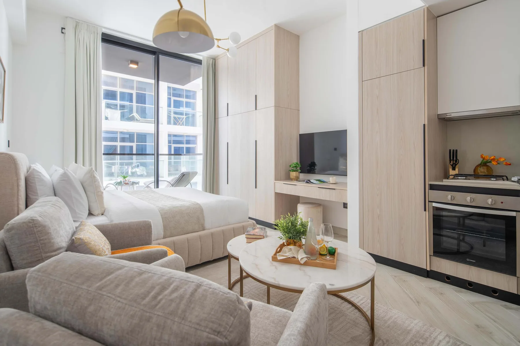 A bright studio living area with a bed, seating, and a compact kitchen.