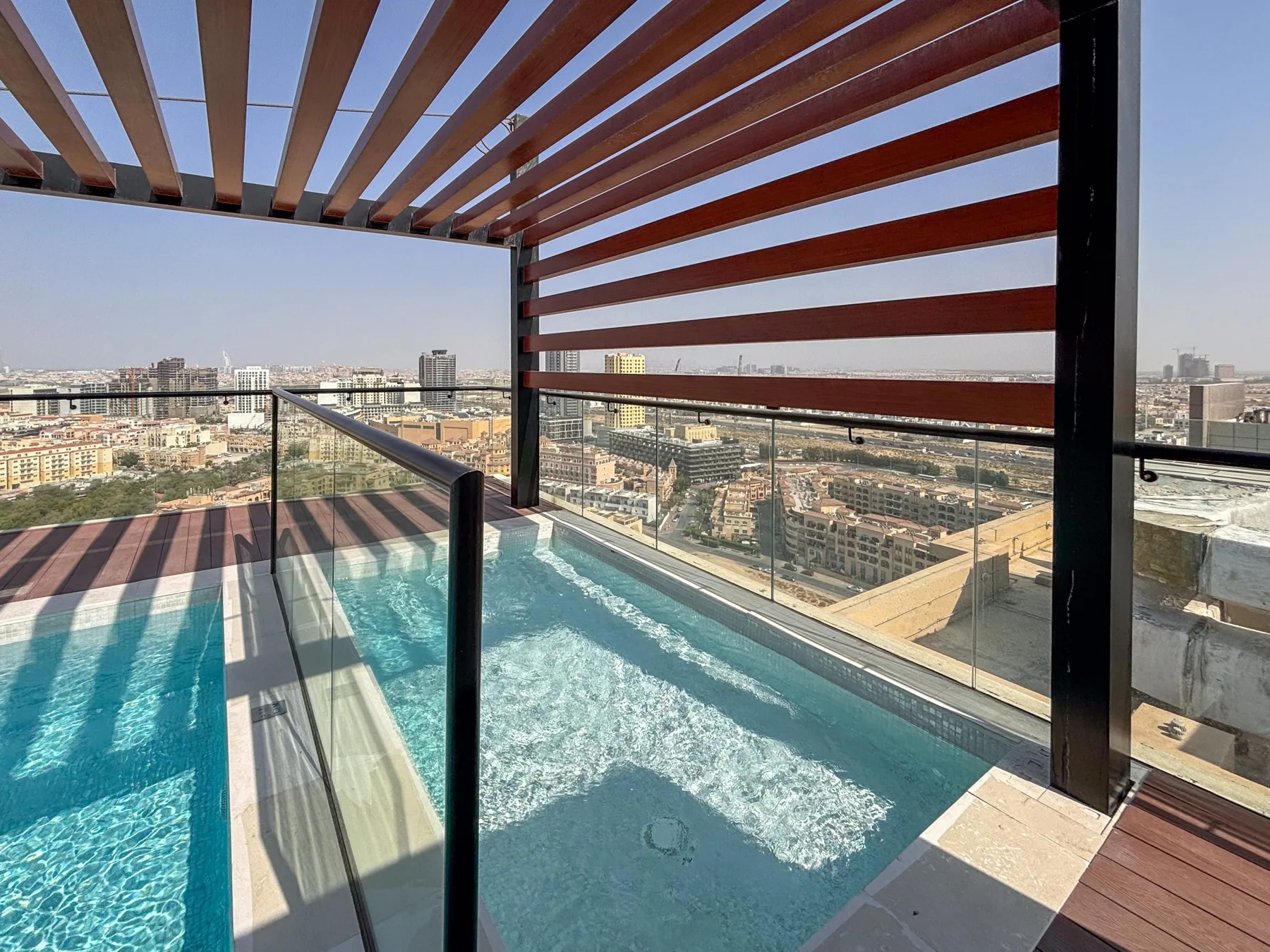 A rooftop pool sits beside glass railings and a slatted shade structure, overlooking the city.