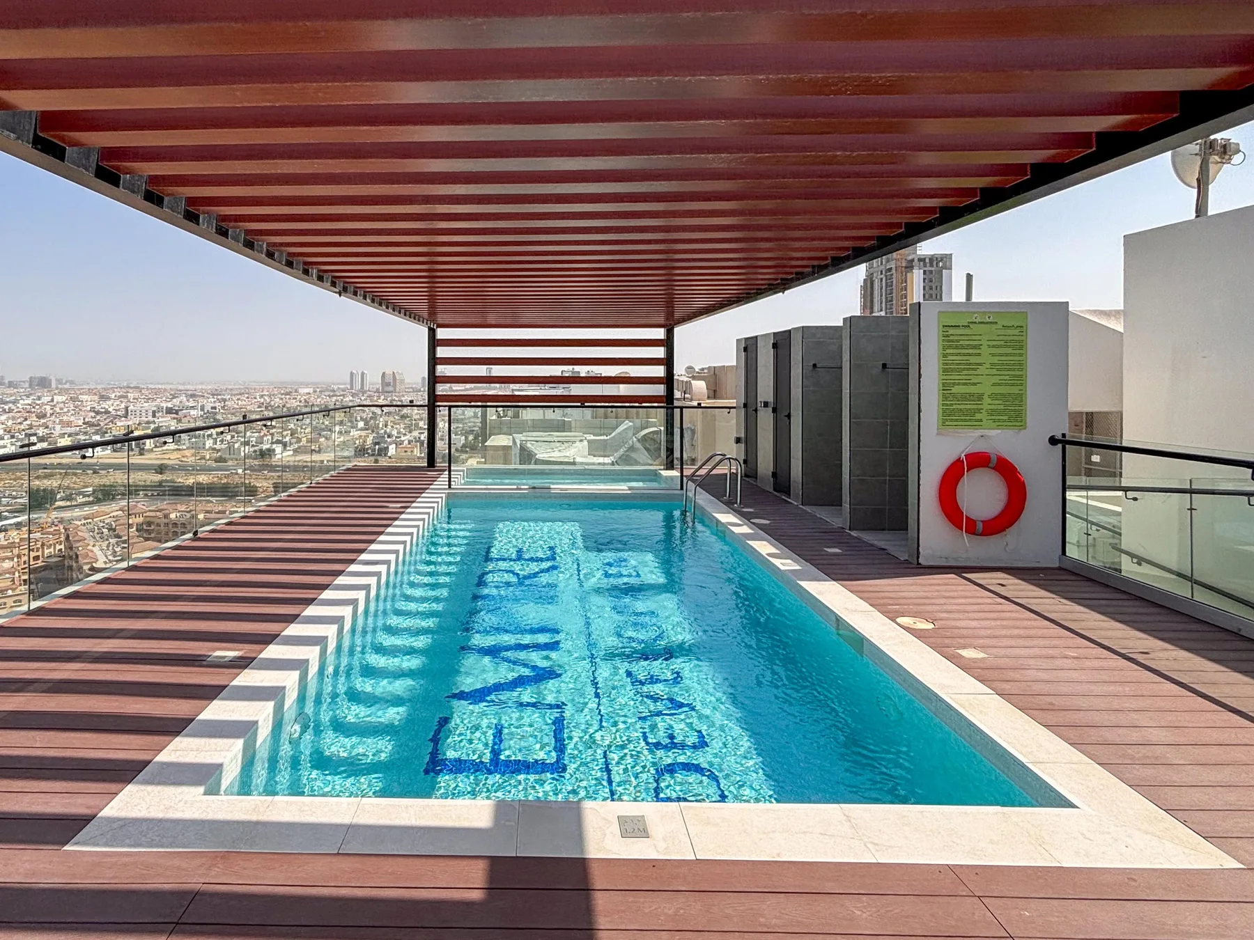 A rooftop swimming pool sits beneath a slatted canopy, with city views beyond.