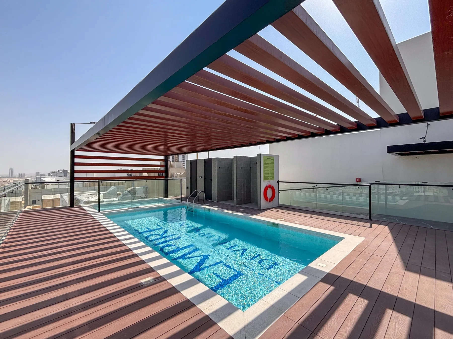 A rooftop swimming pool sits beneath a slatted pergola beside a glass railing.