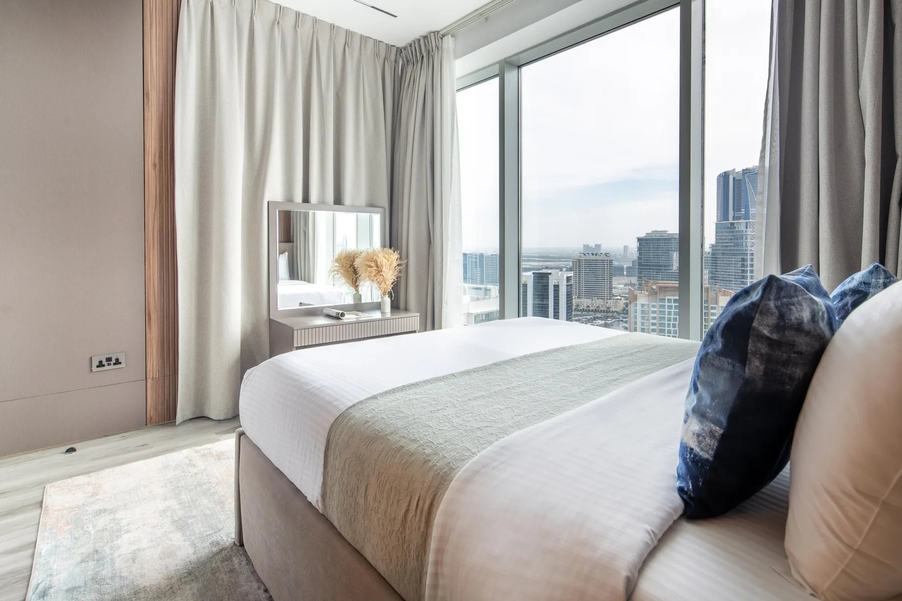 Un dormitorio con una cama bien tendida junto a grandes ventanales con vistas a edificios de la ciudad.
