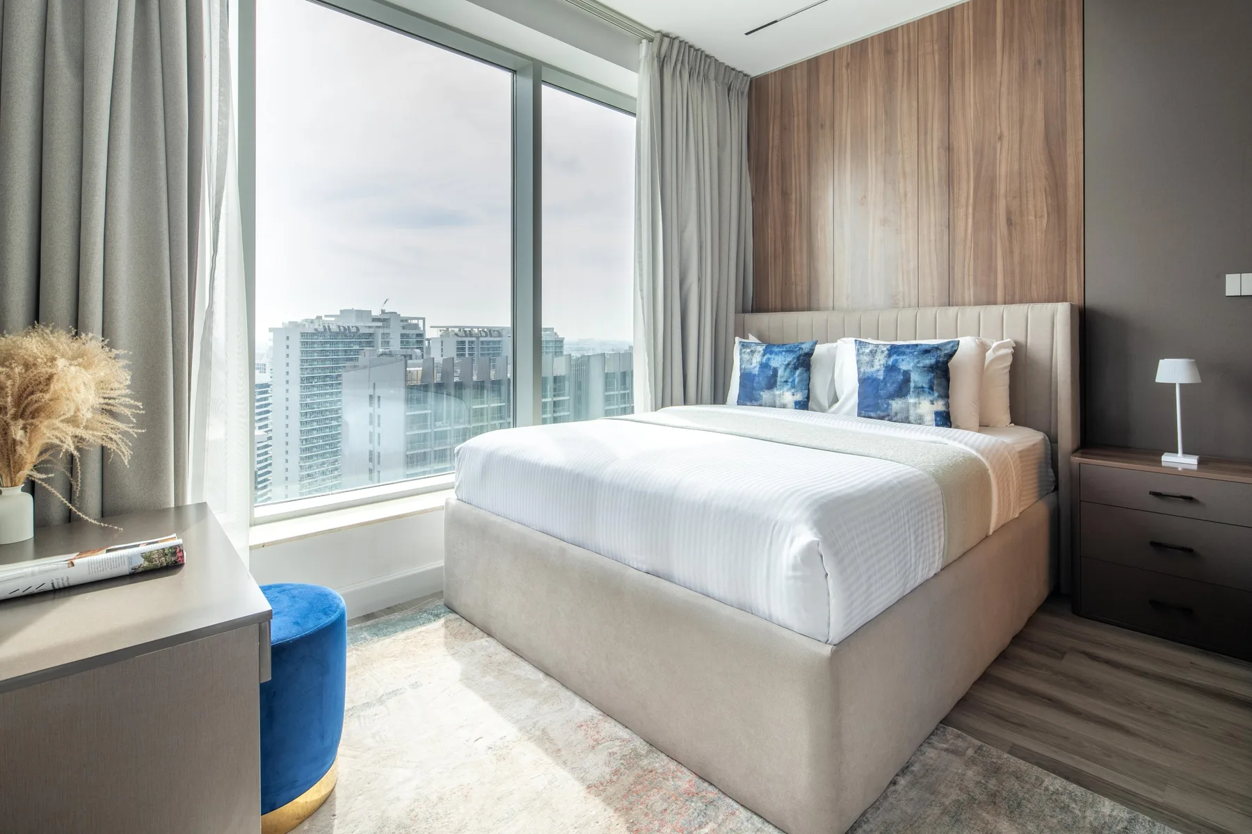Un dormitorio luminoso con una cama grande junto a ventanales con vistas a edificios de la ciudad.