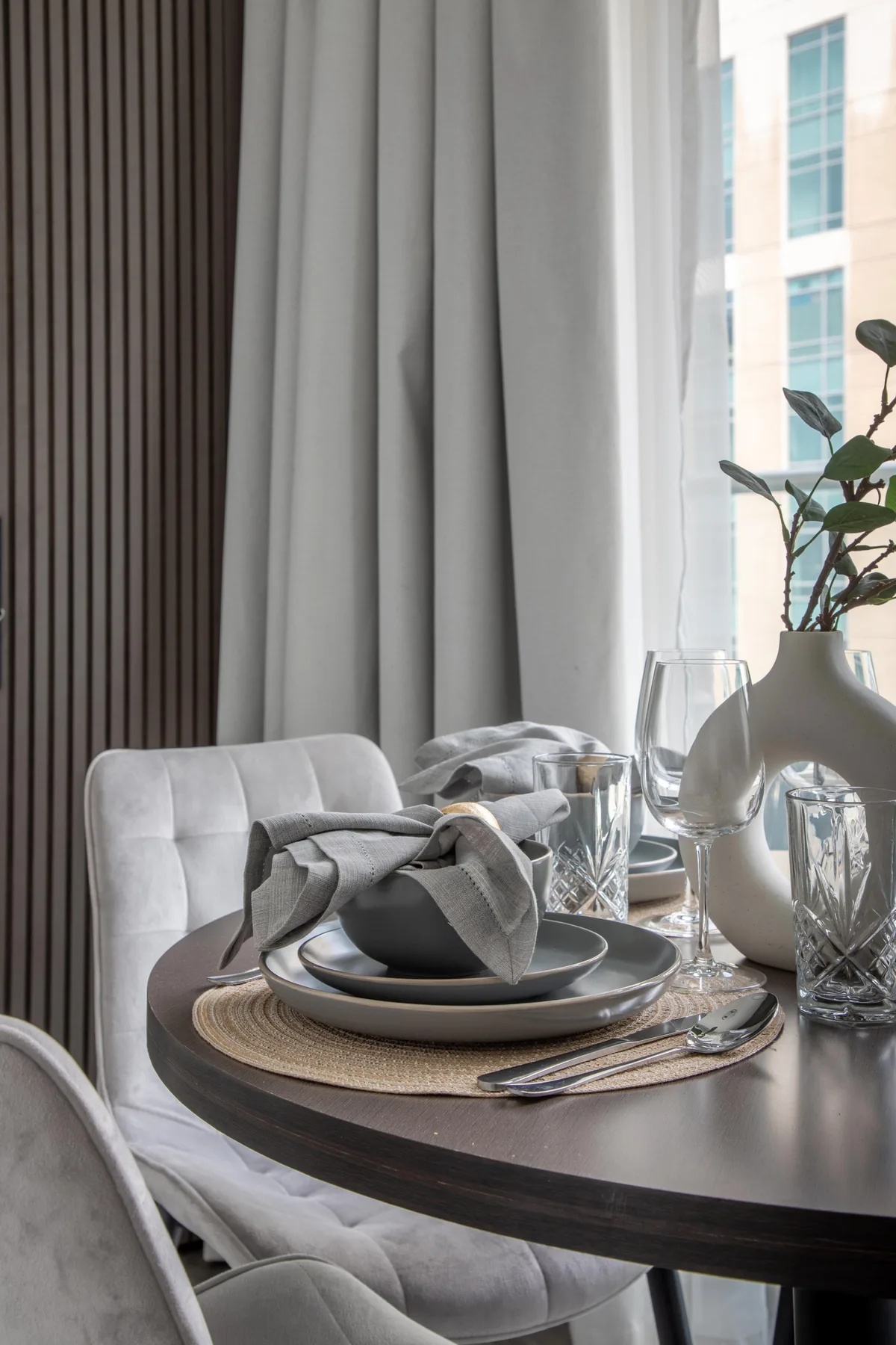 Una mesa de comedor está puesta con platos y vasos junto a una ventana luminosa.