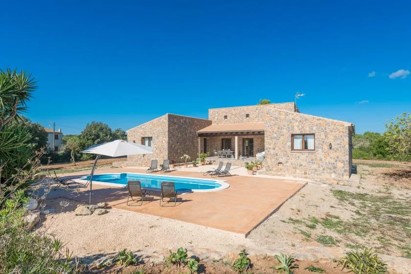 A stone house overlooks a swimming pool and paved patio in a sunny outdoor setting.