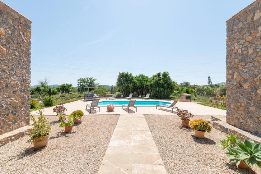 An outdoor swimming pool sits beyond a paved path, surrounded by gravel and plants.
