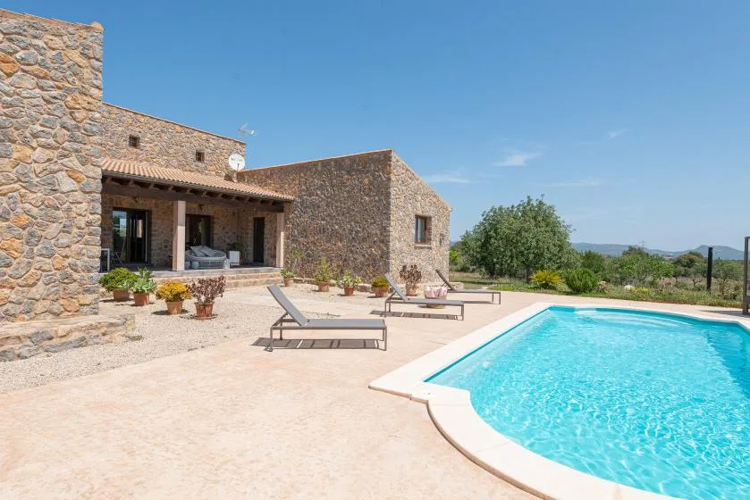 A stone house opens onto a sunny poolside terrace with outdoor seating.