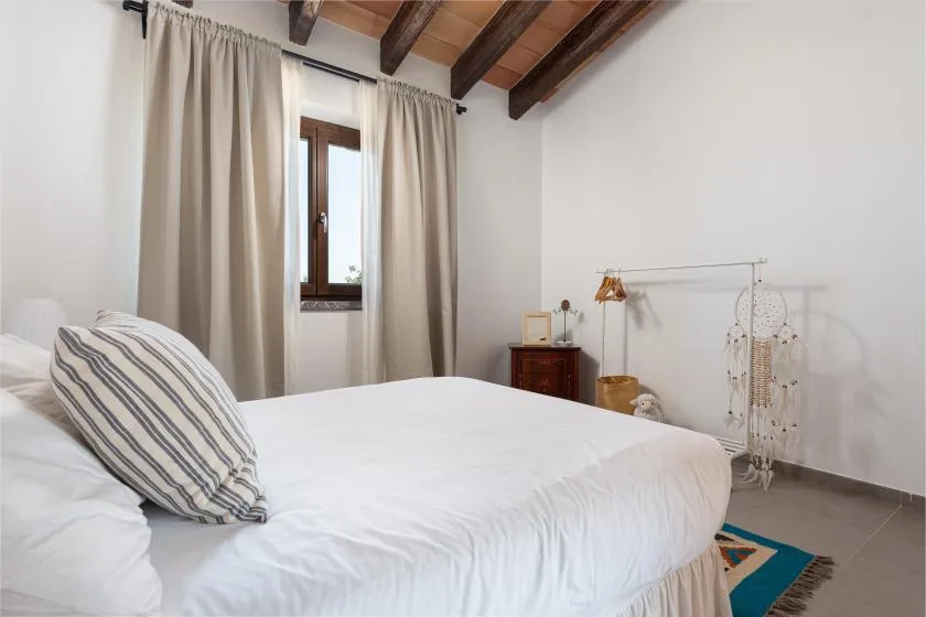 A bedroom with a bed, curtained window, and exposed wooden ceiling beams.