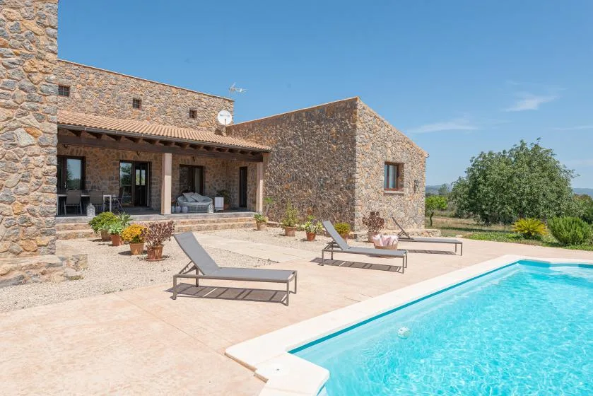 A stone house opens onto a sunny patio with a swimming pool and lounge seating.