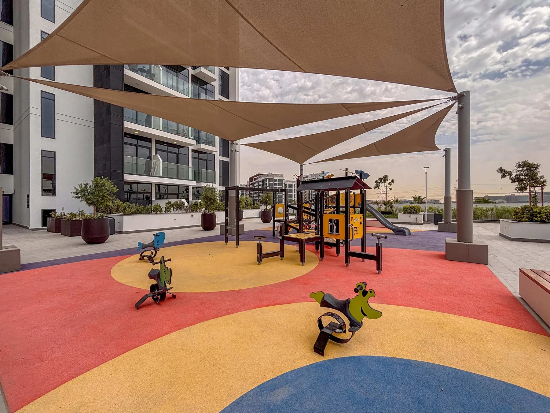Un colorido parque infantil al aire libre está bajo toldos de sombra junto a un edificio moderno.