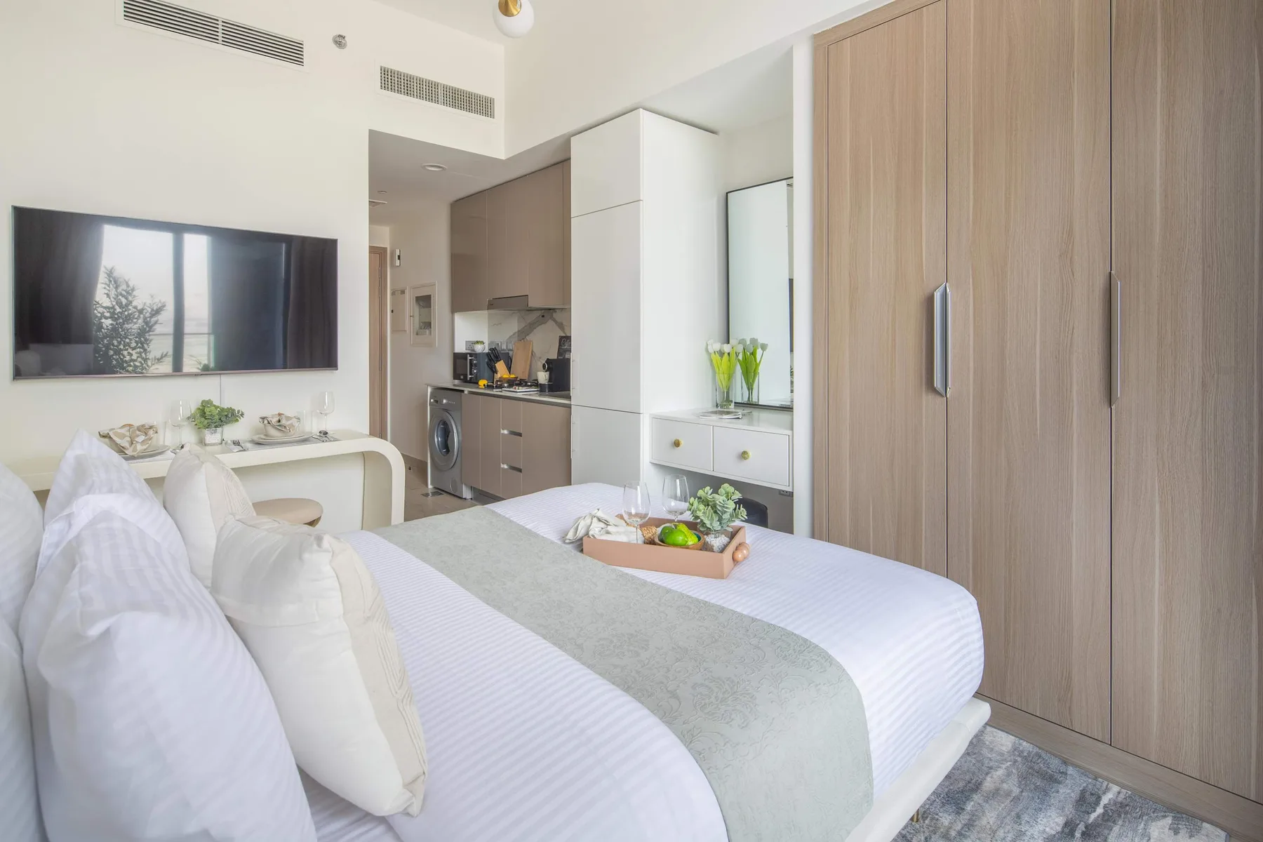 Un dormitorio luminoso con ropa de cama blanca, un televisor en la pared y una pequeña cocina.