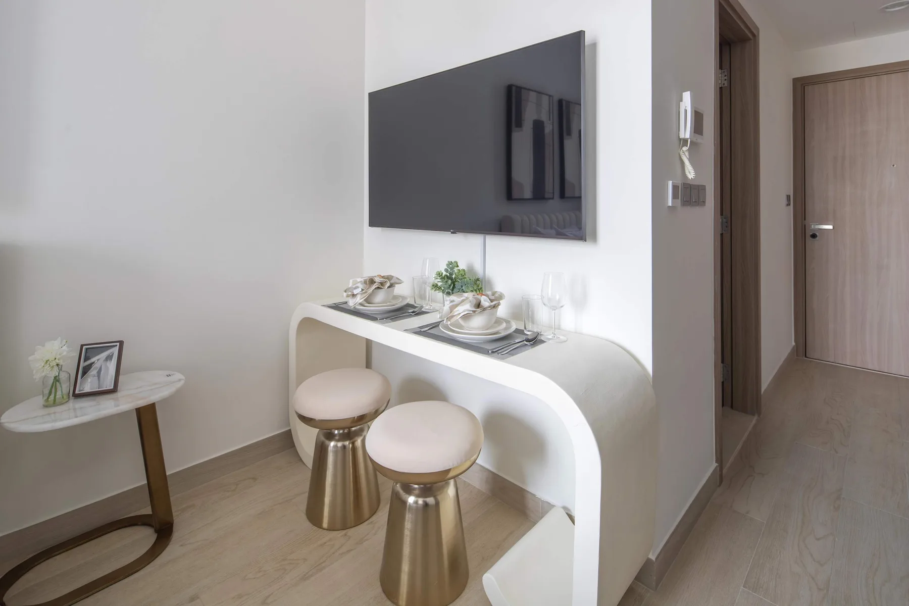 Un rincón de comedor con una mesa blanca curva, dos taburetes y un televisor en la pared.