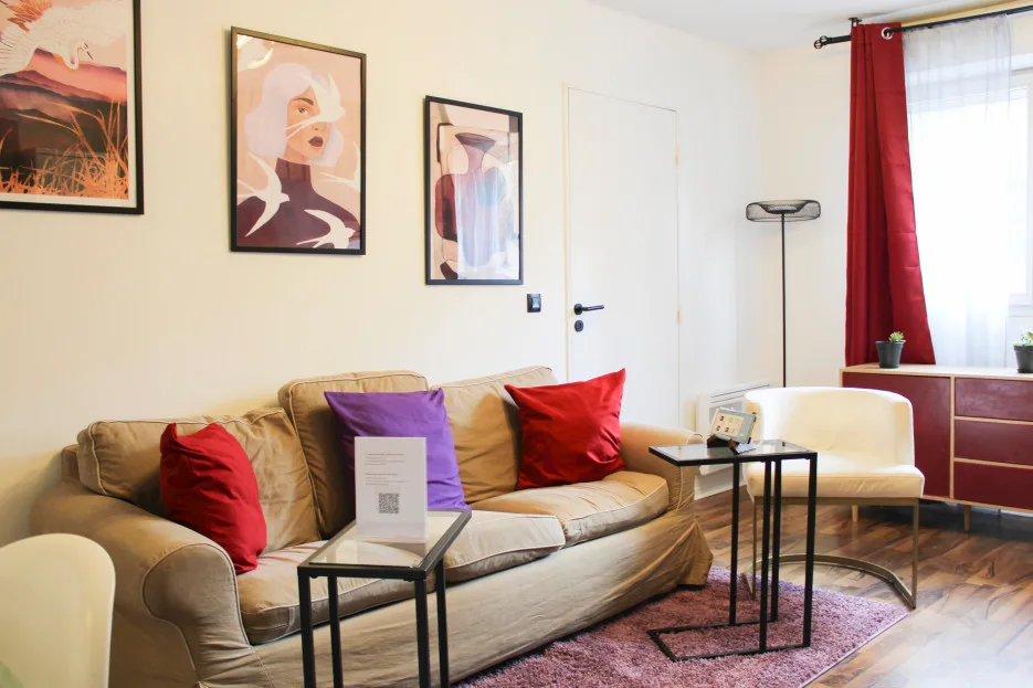A living room with a tan sofa, colorful artwork, and a red curtain by the window.