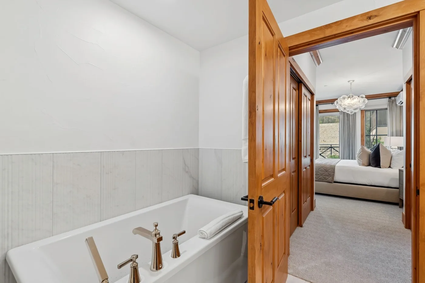A bathroom bathtub sits beside an open wooden doorway leading to a bedroom.