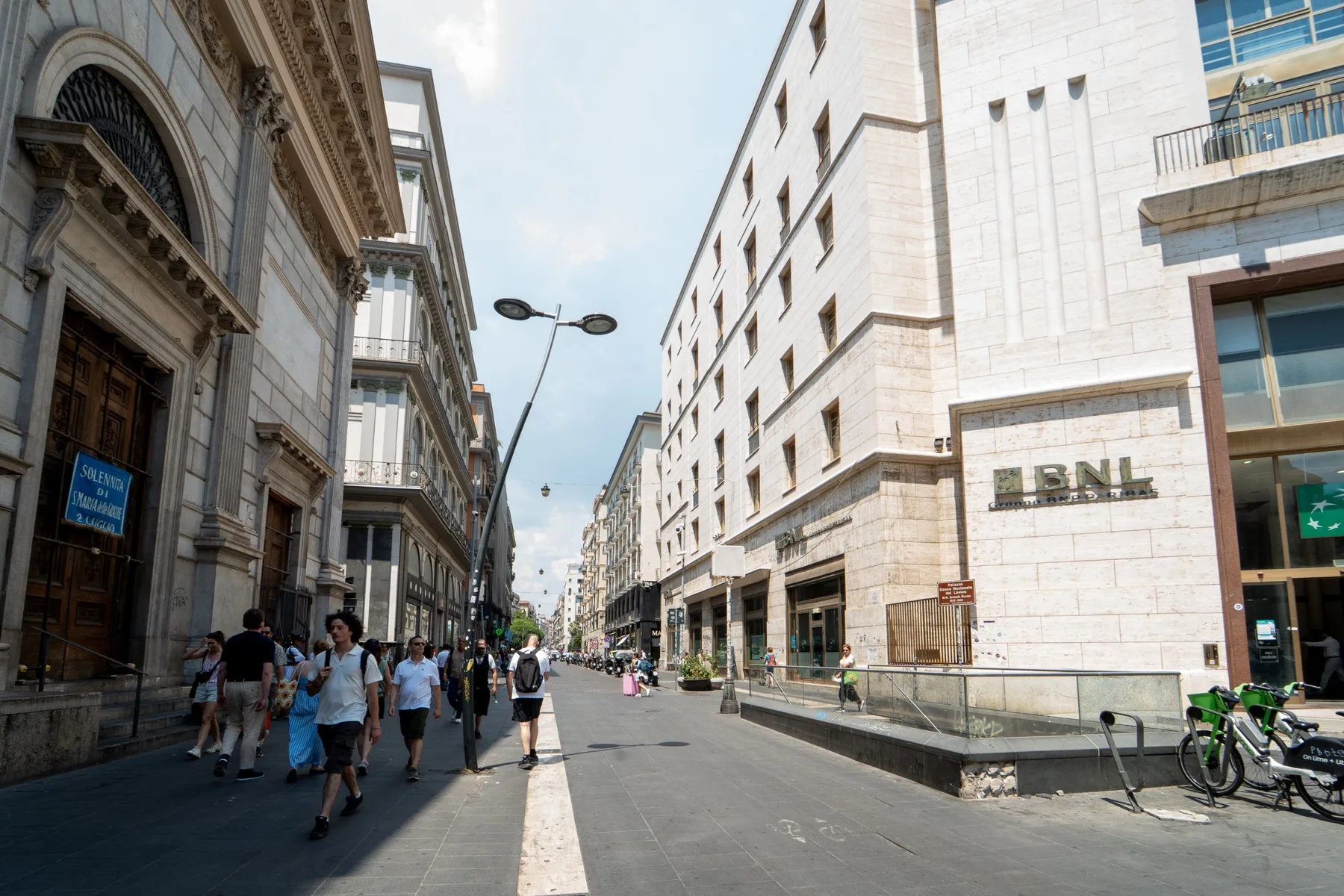 Una calle peatonal transcurre entre edificios altos de piedra, con personas caminando por las aceras.