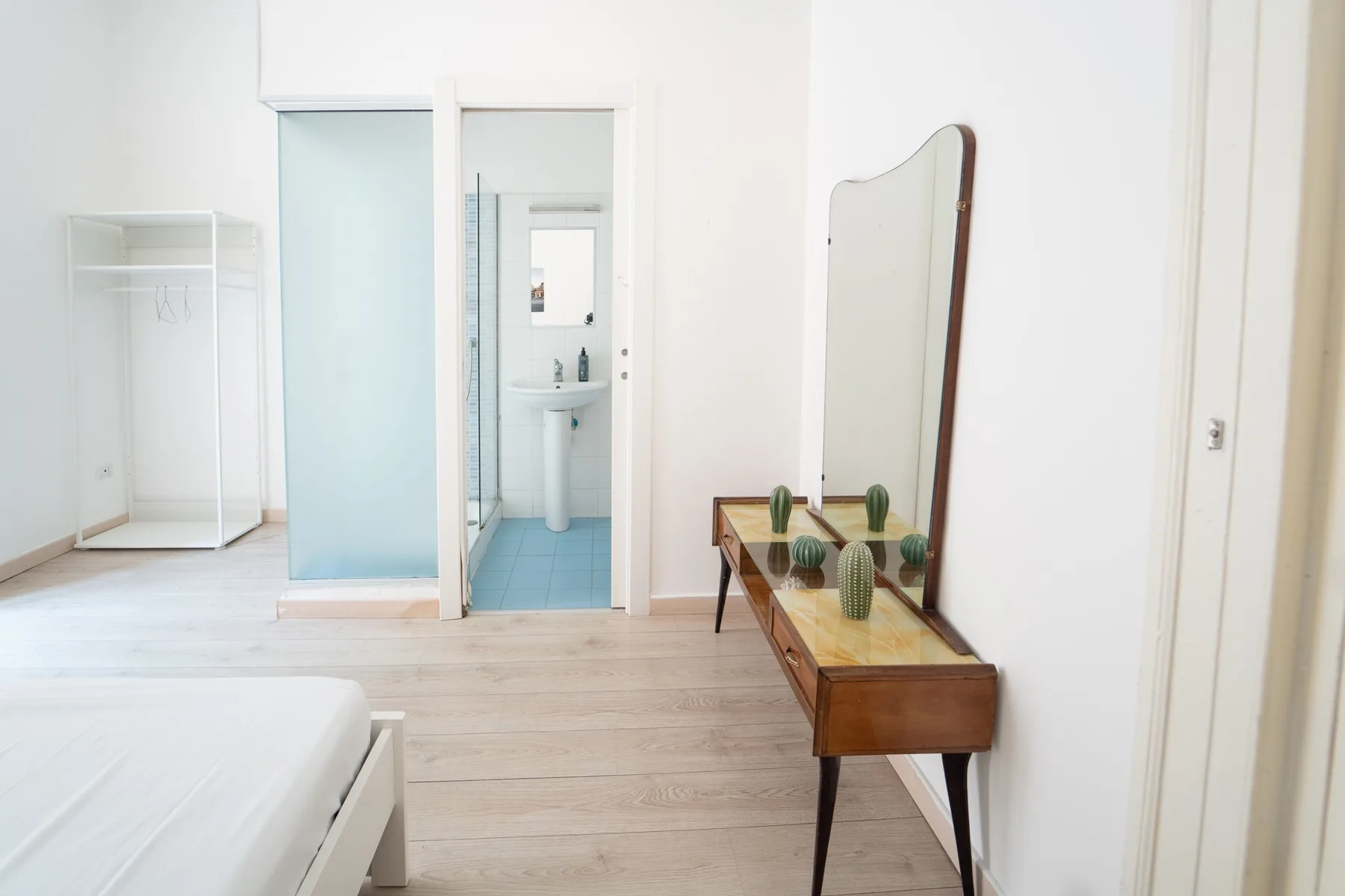 Un dormitorio luminoso con una cama blanca, una consola de madera y un baño contiguo.