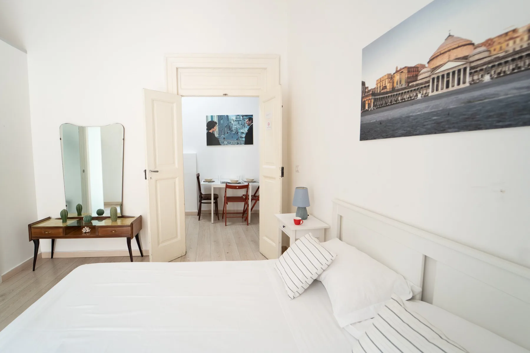 Un dormitorio luminoso con una cama blanca y una puerta abierta hacia un comedor.