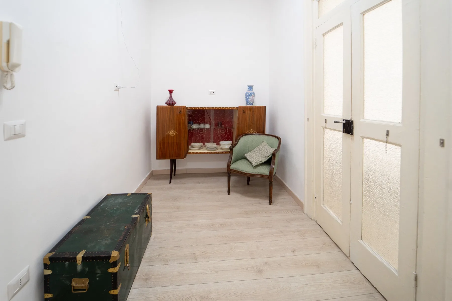 Una habitación luminosa con una vitrina de madera, una silla y un baúl oscuro.