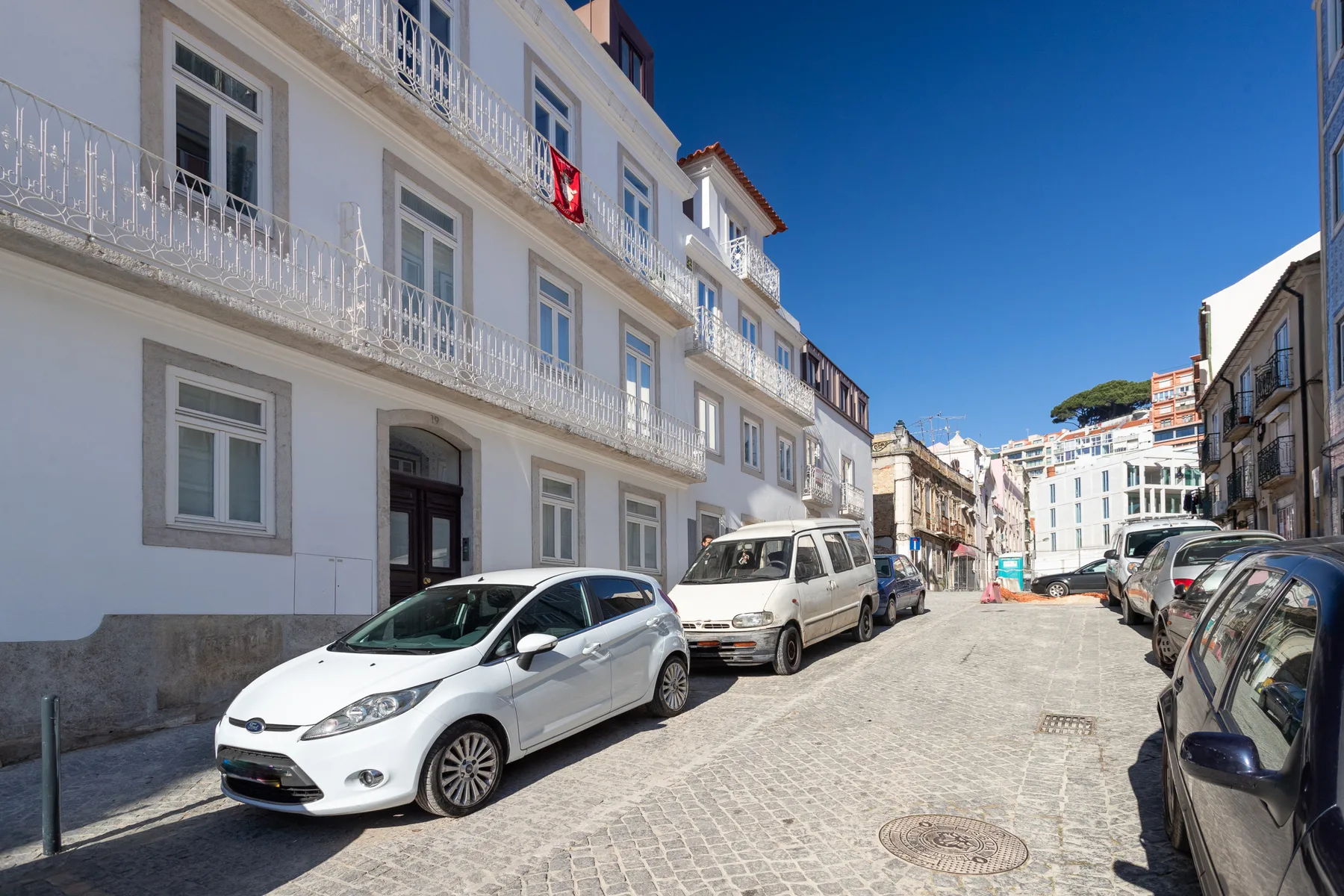 A cobblestone street lined with parked cars and white buildings.