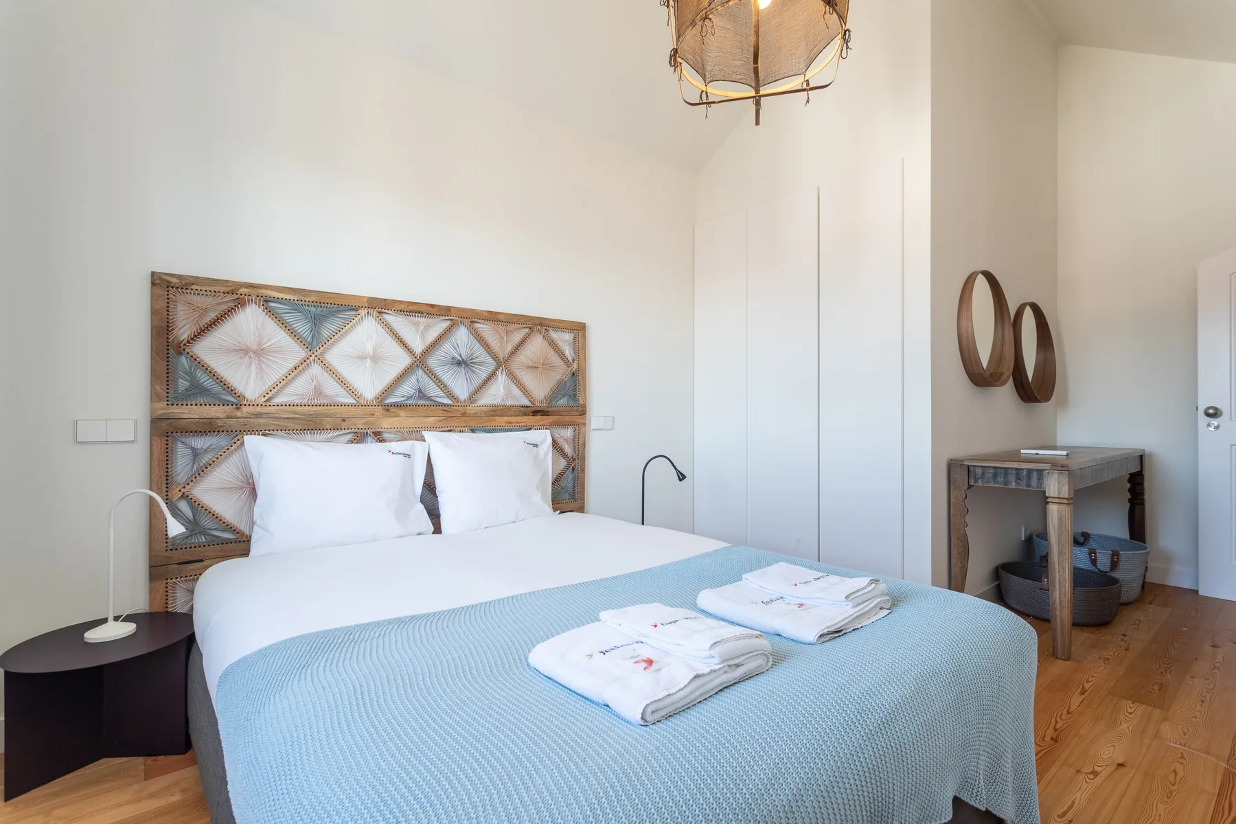 A bright bedroom with a double bed, patterned headboard, and folded towels.