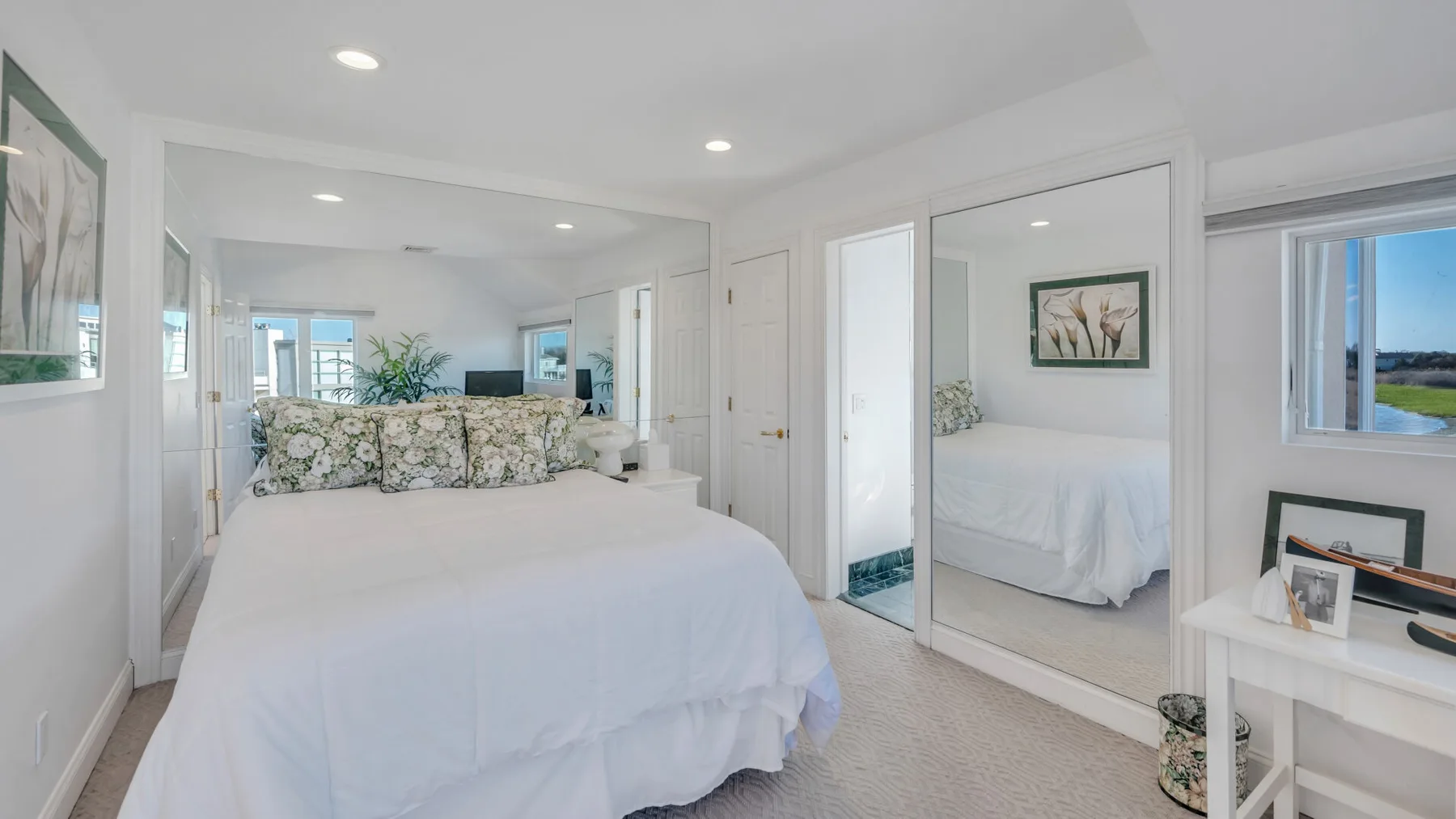 Un dormitorio luminoso con una cama blanca, puertas de armario con espejos y una ventana.