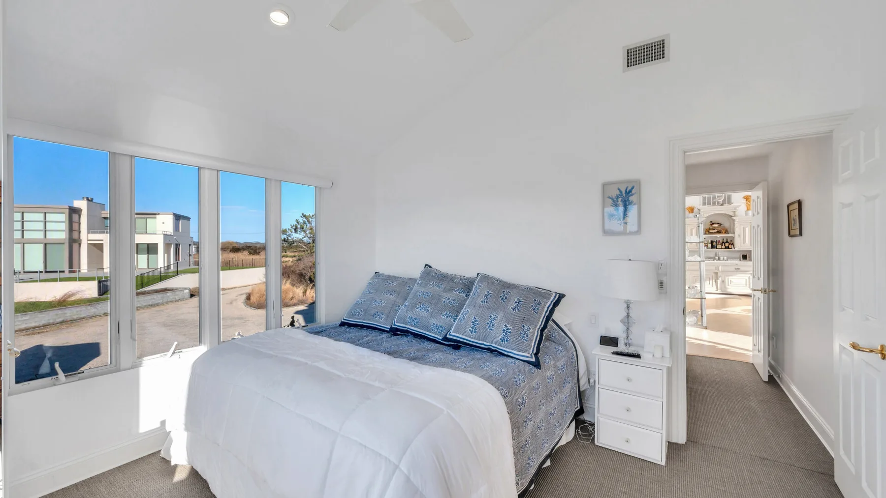Un dormitorio luminoso con una cama junto a grandes ventanas que dan a edificios cercanos.