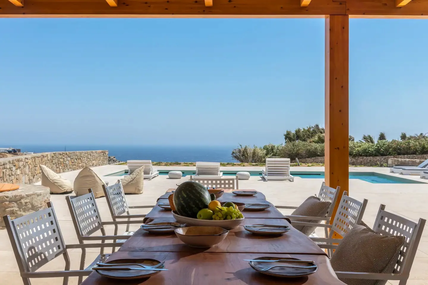 An outdoor dining table sits beneath a wooden pergola beside a pool overlooking the sea.