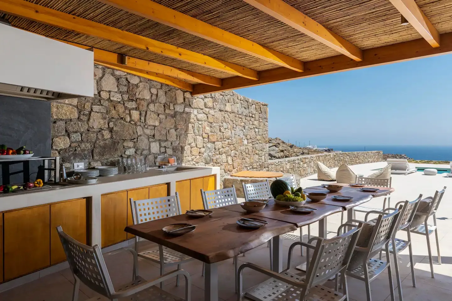 A covered outdoor dining area overlooks the sea, with a long table beside an outdoor kitchen.