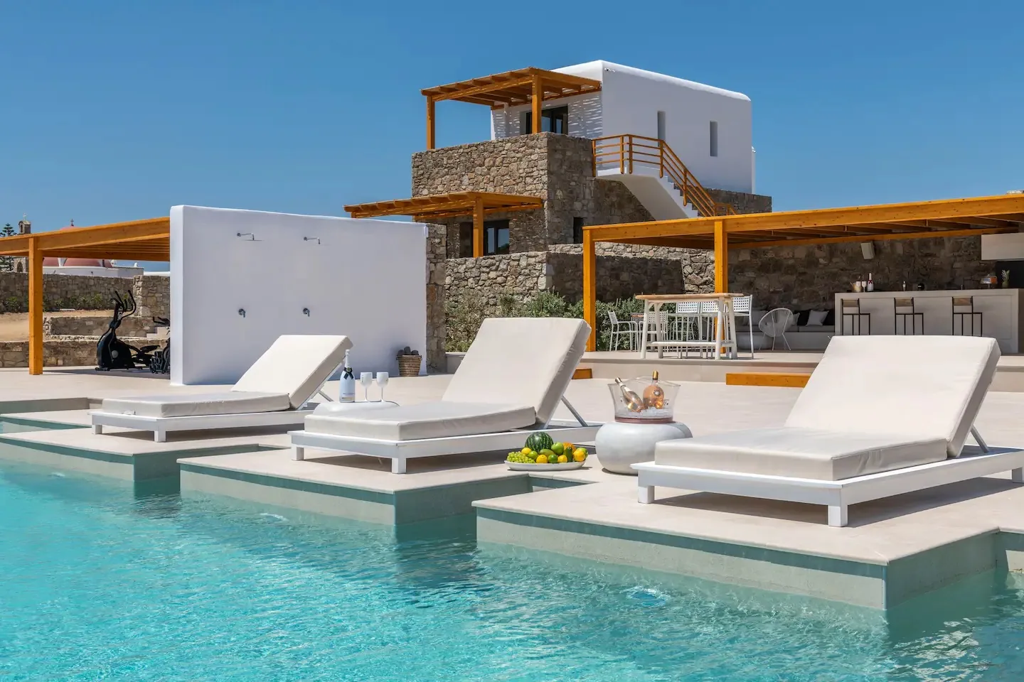 A swimming pool with white loungers sits beside a modern stone-and-white villa.