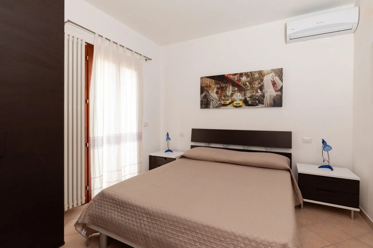 Un dormitorio con una cama entre dos mesitas de noche y una ventana luminosa con cortinas.